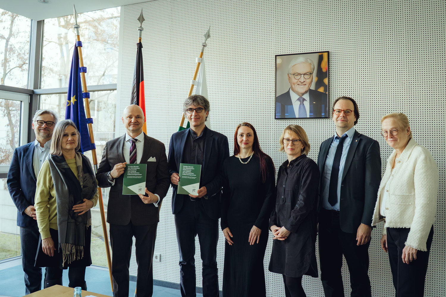 Acht Personen stehen mit Blick zum Betrachter. Zwei Herren halten jeweils eine Broschüre präsentierend in ihren Händen.